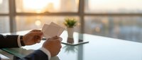 Professional businessman hands holding blank white business card at modern office desk during golden hour sunset with city view background.