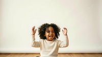 Young child with dark curly hair and medium skin tone wearing cream-colored long-sleeve shirt, arms raised in joyful celebration against neutral background.