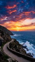 Dramatic sunset over coastal cliffside road with golden and orange sky, crashing ocean waves, and winding mountain path. Perfect for travel, nature, and landscape design projects.