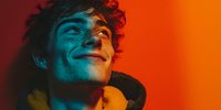Young man with curly hair smiling under dramatic red and blue lighting, wearing yellow hoodie against vibrant orange background.