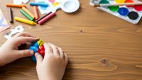 Child hands working with colorful modeling clay on wooden table surrounded by art supplies including watercolor paints and crayons for creative learning activities.
