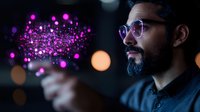 Young bearded South Asian man wearing glasses interacts with glowing purple digital particles floating in dark space, creating futuristic technology visualization effect.