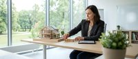 Female architect or real estate professional with dark hair and South Asian ethnicity, wearing formal black blazer, presenting wooden architectural model at modern office desk with large windows overlooking green landscape, planning residential project.