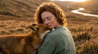 Young woman with curly hair embracing red fox in golden sunset light over highland landscape with river, wildlife conservation and nature connection concept.