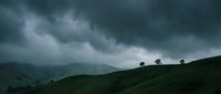 Dramatic storm clouds gathering over rolling green hills with silhouetted trees creating moody atmospheric landscape for weather and nature concepts.