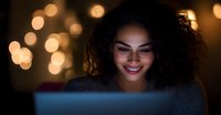 Young African American woman smiling while looking at laptop screen in dark room with warm bokeh lights creating cozy atmosphere, night work from home concept.