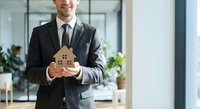 Professional businessman in suit holding wooden house model in modern office space representing real estate investment and property development services.