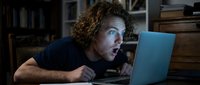 Man with curly brown hair and light skin, wearing dark clothing, focused intently on laptop screen in dimly lit home office workspace for remote work and concentration themes.