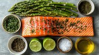Fresh salmon fillet with herbs, spices, asparagus, lime halves, and olive oil on gray background, ready for cooking healthy meal.