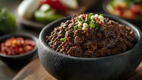 Homemade black bean dip garnished with fresh chopped cilantro and chili in rustic stone bowl, served with salsa and fresh vegetables on wooden table.