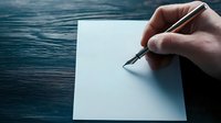 Male hand holding fountain pen writing on blank white paper against dark wooden table surface in moody lighting, capturing moment of creative inspiration.