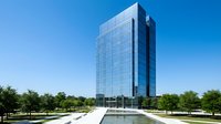 Modern glass office tower with reflective facade rising against clear blue sky, surrounded by green landscaping and reflecting pool, suitable for corporate, real estate, and business concepts.
