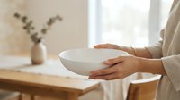 Close-up of hands holding empty white ceramic bowl in bright kitchen with wooden table and dried flowers in vase, suitable for food, cooking, and lifestyle content.