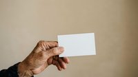 Senior male hand holding blank white business card against neutral beige background for corporate branding and professional networking purposes.