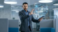 Young male professional, Caucasian ethnicity, wearing dark business suit and blue shirt, holding laptop while interacting with holographic digital interface displaying security and technology icons in modern office environment.