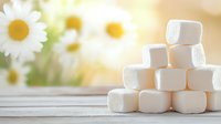 White fluffy marshmallows stacked in pyramid shape on wooden surface with soft focus chamomile flowers and natural sunlight in background creating cozy atmosphere.