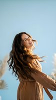 Happy young woman with flowing brown hair laughing joyfully in brown dress against blue sky background for lifestyle and wellness concepts.