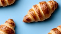 Fresh baked golden croissants with flaky layered texture and glossy surface arranged on blue background, food photography for bakery and cafe menu.