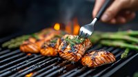 Grilled salmon fillets with herbs and asparagus cooking on barbecue grill with flames. Fork adding seasoning to juicy fish steaks. Close up view of seafood preparation.