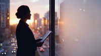Professional businesswoman silhouette using tablet by office window at sunset with city skyline view for corporate technology and urban lifestyle concepts.