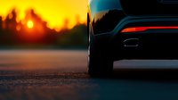 Close-up view of modern car tire and exhaust on asphalt road during dramatic orange sunset with silhouetted forest treeline in background.