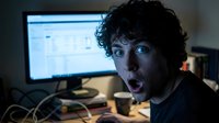 Young man with dark curly hair and South Asian appearance, wearing black shirt, expressing shock or surprise while working at computer desk with monitor displaying data, suitable for tech, business, and online content.