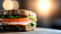 Fresh salmon sandwich with lettuce and tomato on dark rye bread, close-up view against blurred warm lights background, appetizing lunch meal.