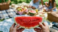 Fresh watermelon slice held in hands at outdoor summer picnic with fruit baskets and checkered blanket in blurred background, ideal for seasonal food and lifestyle content.