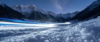 Snowy mountain valley at night with starry sky and blue light trails crossing frozen landscape, suitable for winter backgrounds and nature compositions.