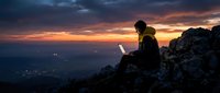 Person working on laptop atop rocky mountain peak during dramatic sunset, silhouetted against golden and purple sky with distant city lights below.