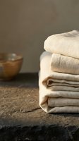 Stack of folded cream linen towels on dark stone surface with blurred wooden bowl, ideal for spa, wellness, hospitality and home decor design projects.