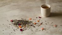 Scattered loose tea leaves with delicate pink and purple flower petals on neutral surface, ceramic cup with fresh brewed tea in soft natural light.