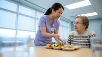 Asian female nurse in purple scrubs serving healthy meal to elderly Caucasian woman patient in modern hospital room with natural lighting for healthcare nutrition.