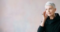 Senior woman with short white hair and fair complexion wearing black clothing, touching her face thoughtfully against neutral background for portrait and lifestyle use.
