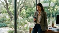 Young woman of mixed ethnicity in casual business attire using smartphone while working at desk near large windows overlooking garden landscape. Remote work, modern office.