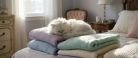 White fluffy cat resting on stack of folded pastel blankets in bright bedroom with natural light, home decor and pet lifestyle.