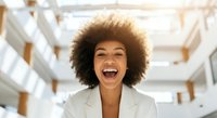Young woman with medium brown skin tone and voluminous curly afro hairstyle wearing beige blazer, smiling broadly with mouth open in modern office environment with white walls and natural lighting.