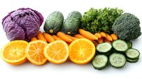 Fresh colorful vegetables and fruits arranged in row on white background, including purple cabbage, zucchini, carrots, orange slices, cucumber, and broccoli.