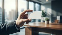 Professional businessman holding blank white business card in modern office space with city view background for corporate branding and networking purposes.