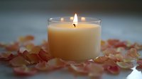 Glowing white candle in glass holder surrounded by scattered pink and peach rose petals creating romantic atmosphere on blurred background. Suitable for spa and wellness.