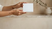 Woman with medium skin tone holding blank white card in both hands against neutral beige background with dried plant decor, suitable for business cards, invitations, and promotional materials.