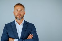 Confident middle-aged Caucasian businessman with gray beard wearing navy blue suit and white shirt, arms crossed, against light gray background. Professional portrait.