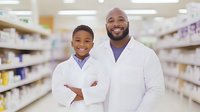 African American pharmacist and young boy in white coats smiling in pharmacy aisle, representing healthcare education, mentorship and future medical professionals.