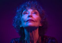 Mature woman with curly hair in dramatic purple and blue lighting gazes upward, wearing elegant jewelry. Artistic portrait capturing confidence and contemplation.