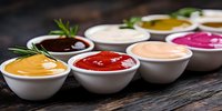 Assorted colorful sauces and dips in white ceramic bowls on rustic wooden background with fresh herbs garnish, panoramic food composition.