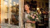 Mature woman with light skin tone and shoulder-length brown hair, wearing a beige apron, holding a fresh flower arrangement with pink roses and white blooms in a rustic flower shop with vintage wooden doors and warm lighting.