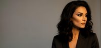 Professional Middle Eastern businesswoman with dark curly hair in black blazer looking confidently away from camera against neutral background.