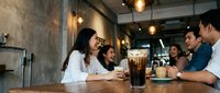 Young diverse professionals enjoying coffee break conversation in modern industrial cafe with warm lighting and concrete walls.