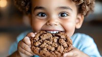 Young child with curly brown hair and light skin tone eating a chocolate chip cookie, smiling with joy and delight. Suitable for family, parenting, nutrition and food content.
