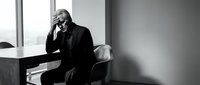 Senior male professional, Caucasian appearance, wearing dark business suit, sitting at desk in modern office, hand to face in contemplative or stressed pose, black and white photography, corporate workplace setting.
